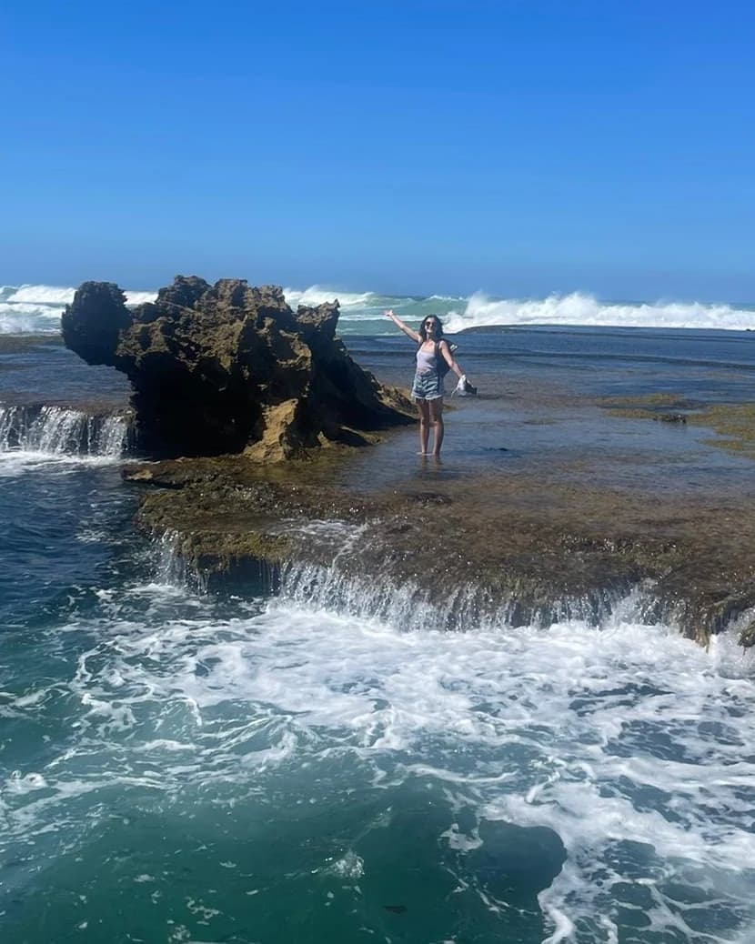 Léa with her arms wide open facing the ocean, next to a rock